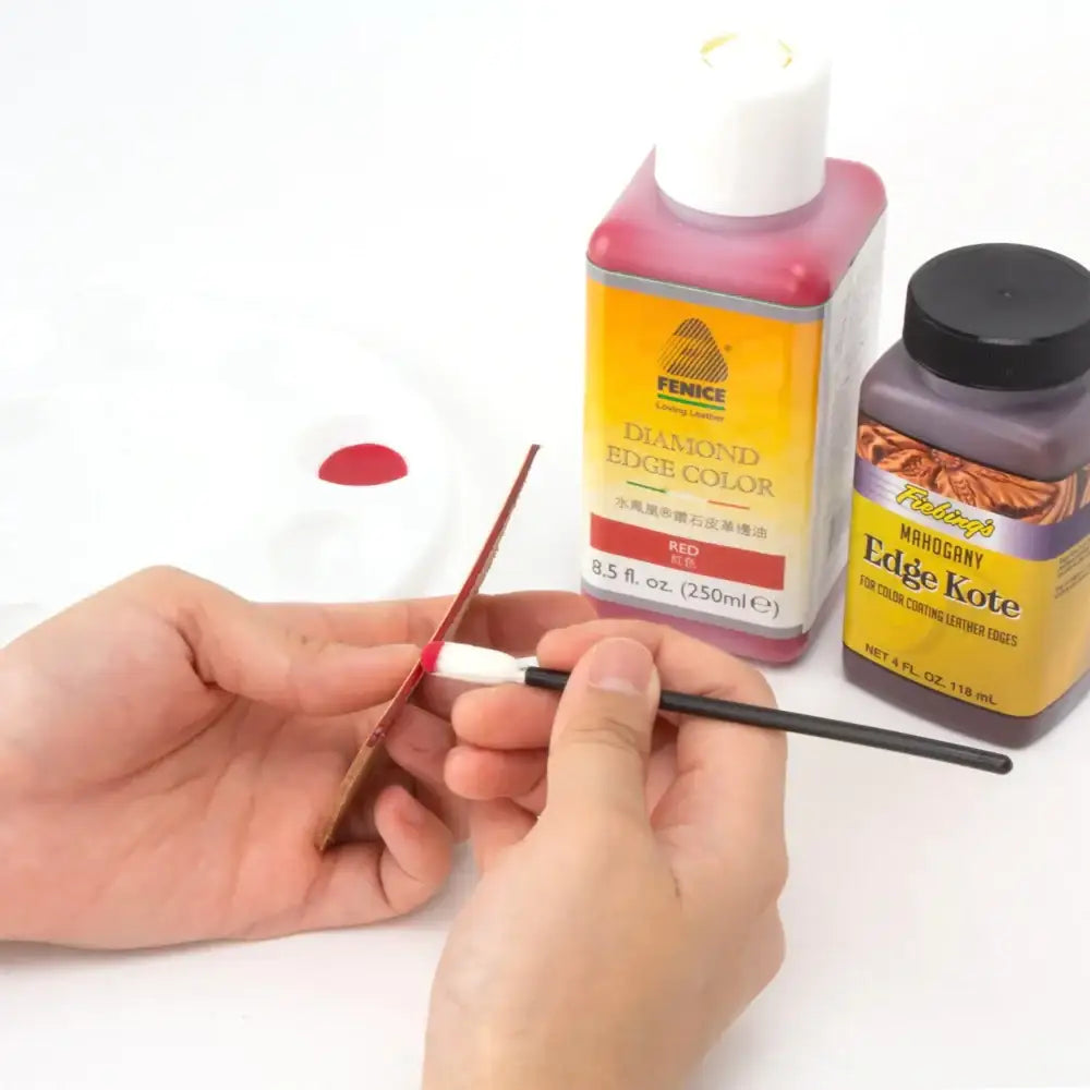 Person trägt rote Kantenfarbe mit Pinsel auf Lederkante auf, Flaschen Fenice Diamond Edge Color und Fiebings Mahogany Edge Kote sichtbar