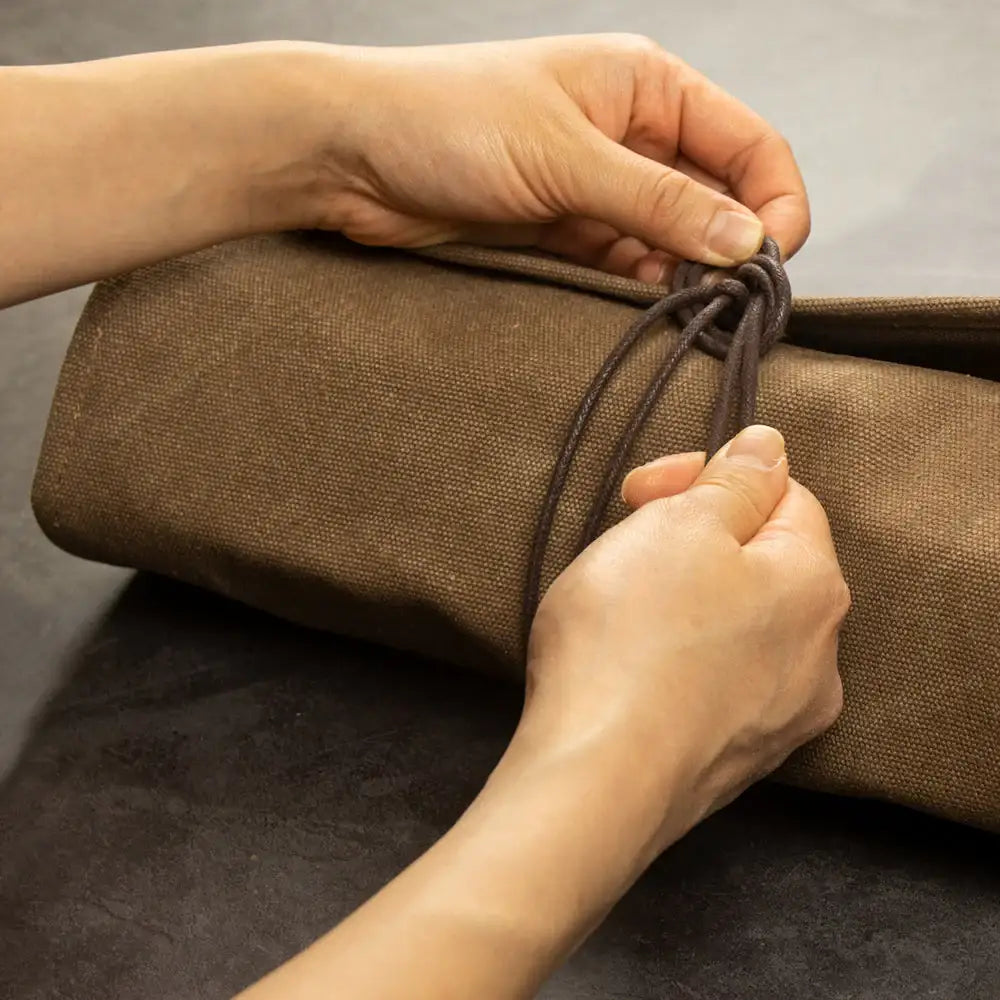 Hände binden braunes Lederband um zusammengerollte Werkzeugtasche auf dunklem Tisch