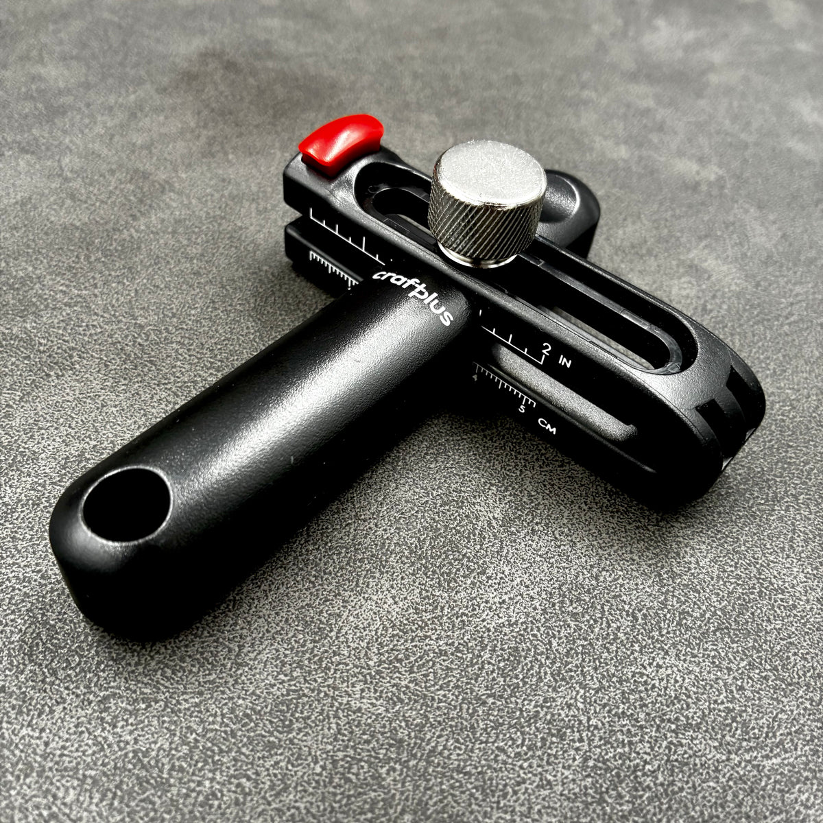 Schwarzer Leder-Prägestempel mit verstellbarer Skala und rotem Hebel auf grauem Untergrund.