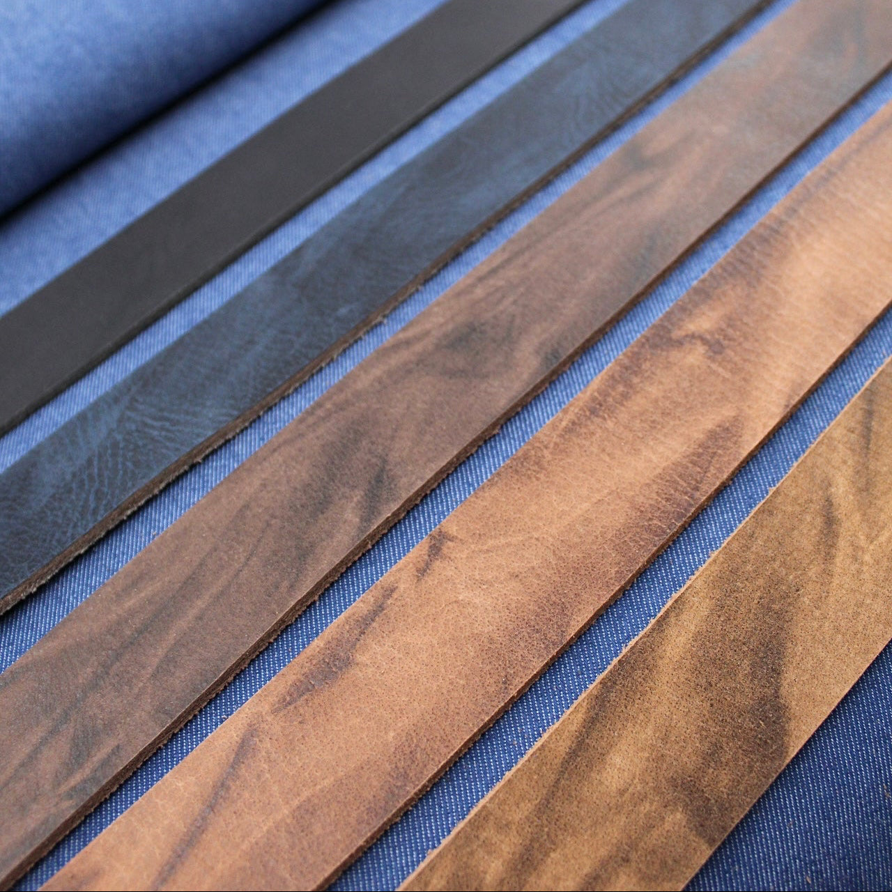 Mehrere Lederstreifen aus Vollbüffelleder in verschiedenen Braun- und Schwarztönen liegen locker geschwungen auf blauem Stoff.