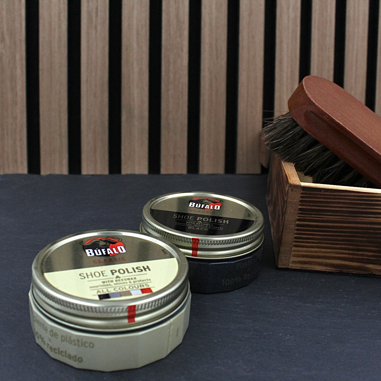 Zwei Dosen Büffalo Schuhcreme und Ledersohlenbürste im Holzbehälter vor gestreiftem Hintergrund