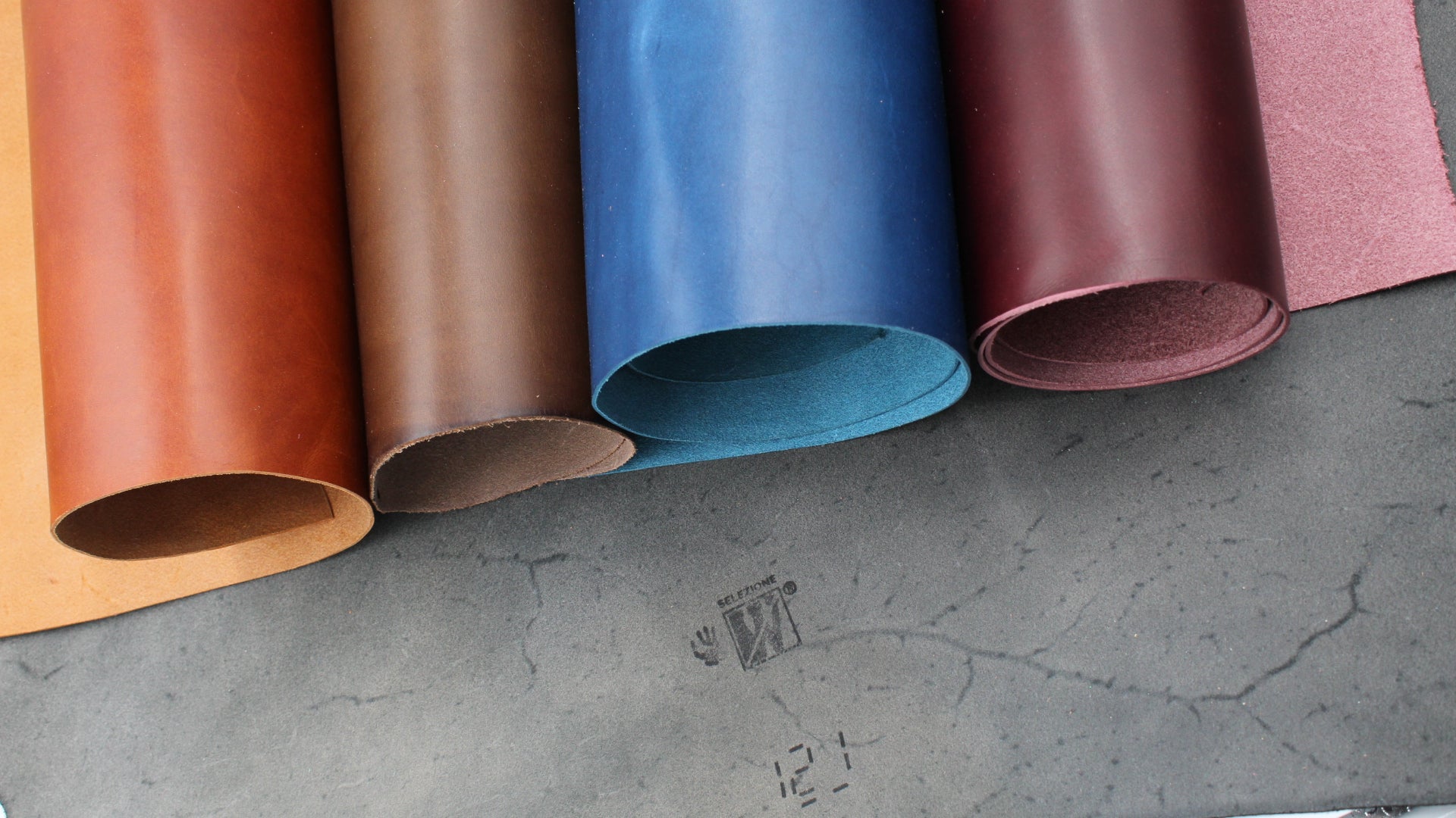 Vier Rollen Walpier Buttero Premium-Leder in den Farben Cognac, Braun, Blau und Weinrot auf grauem Untergrund. Italienisches pflanzlich gegerbtes Vollnarbenleder mit glatter Oberfläche und sichtbarem Walpier-Stempel.